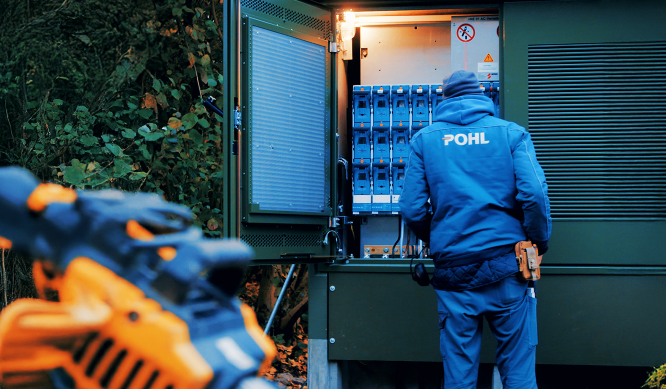 Ein Monteur in blauer POHL-Jacke arbeitet an einem großen grünen Schaltschrank im Freien. Das Innere mit vielen Schaltern und Kabeln ist beleuchtet.