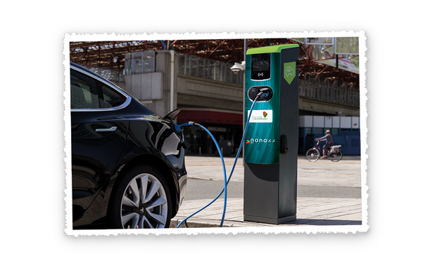 Auto lädt an einer öffentlichen Ladesäule, das Ladekabel steckt, im Hintergrund eine große Halle und vorbeifahrende Personen.