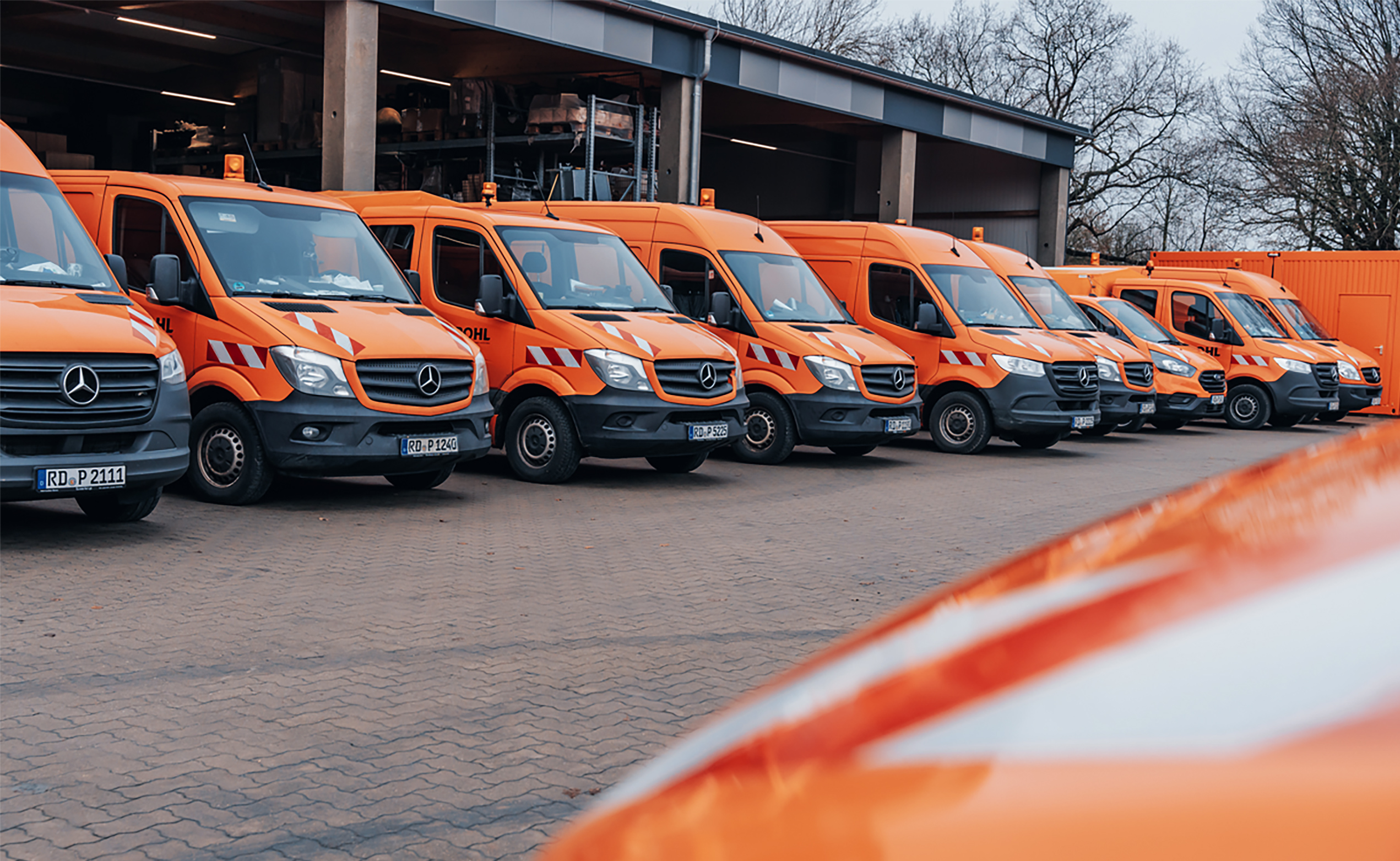 In der Mitte stehen mehrere orangefarbene POHL-Fahrzeuge in einer Reihe auf einem Hof. Links im Bild sind die Fronten der Transporter mit weißen Warnstreifen zu sehen. Rechts im Bild setzt sich die Fahrzeugreihe bis zu einem orangefarbenen Container fort. Im Hintergrund steht eine offene Halle. Vorne im Bild ist eine angeschnittene orangefarbene Motorhaube.