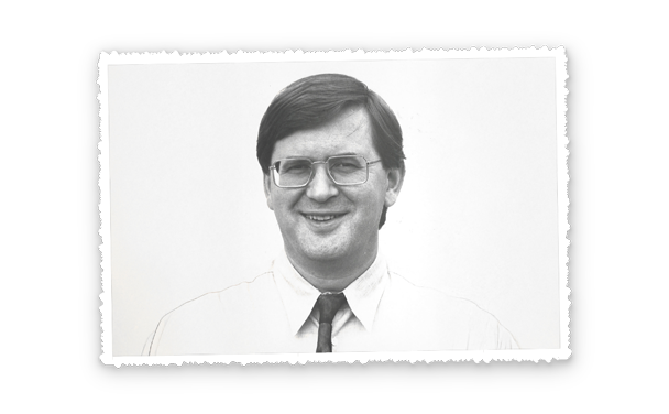 Porträt eines Mannes mit Brille, Hemd und Krawatte, Oberkörper frontal, neutraler heller Hintergrund.