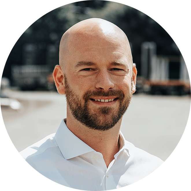 In der Mitte steht eine männliche Person mit kahlrasiertem Kopf und kurzem Bart. Sie trägt ein weißes Hemd; im Hintergrund verläuft eine Straße mit unscharfen, orangefarbenen POHL-Fahrzeugen.