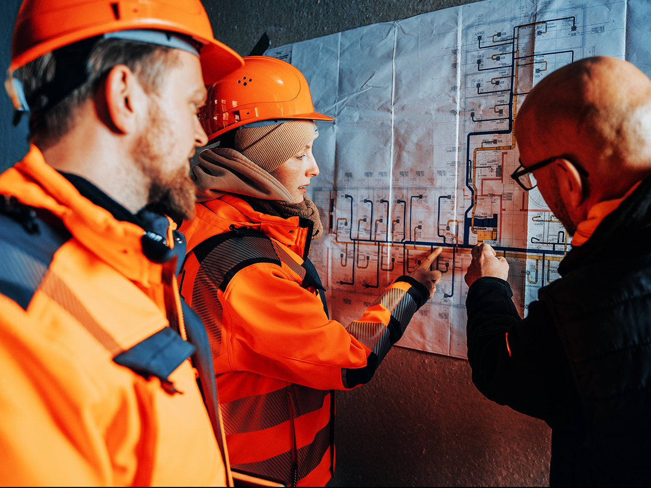 Drei POHL Mitarbeiter mit Schutzhelmen und Arbeitskleidung besprechen Baupläne an einer Wand.