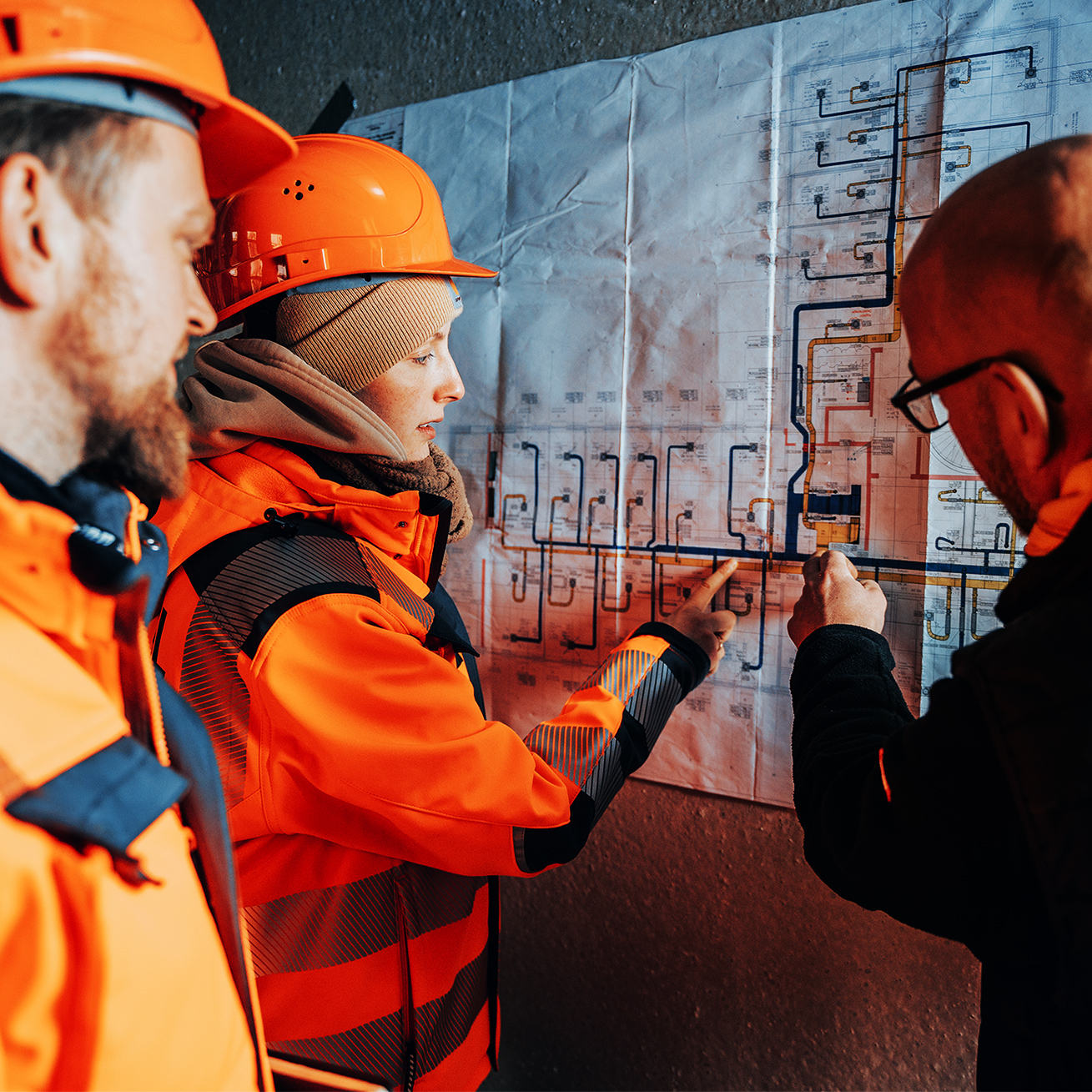 Drei POHL Mitarbeiter mit Schutzhelmen und Arbeitskleidung besprechen Baupläne an einer Wand.