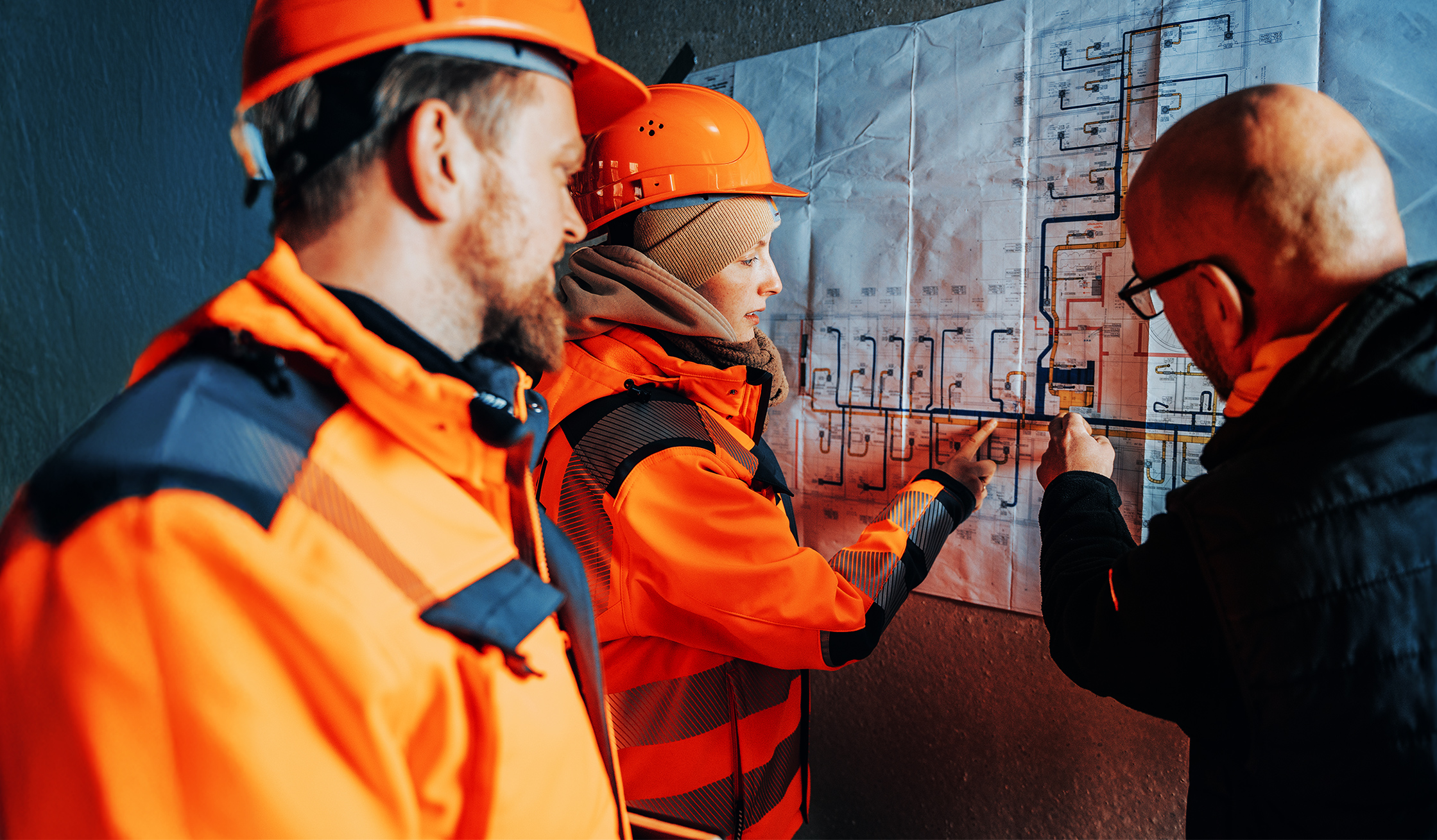 Drei POHL Mitarbeiter mit Schutzhelmen und Arbeitskleidung besprechen Baupläne an einer Wand.