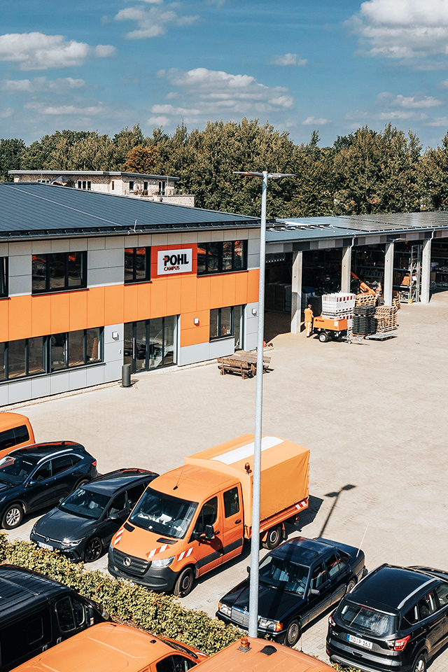 In der Mitte steht ein zweigeschossiges Gebäude mit grauen Fassaden und einem orangefarbenen Band; über dem Eingang hängt das Schild „POHL Campus“. Vorne im Bild parken mehrere orange POHL-Transporter zwischen dunklen Pkw; rechts schließt sich eine lange, offene Lagerhalle mit Regalen und Material an. Im Hintergrund verläuft eine Baumreihe vor blauem Himmel; links sind Nachbargebäude zu sehen.