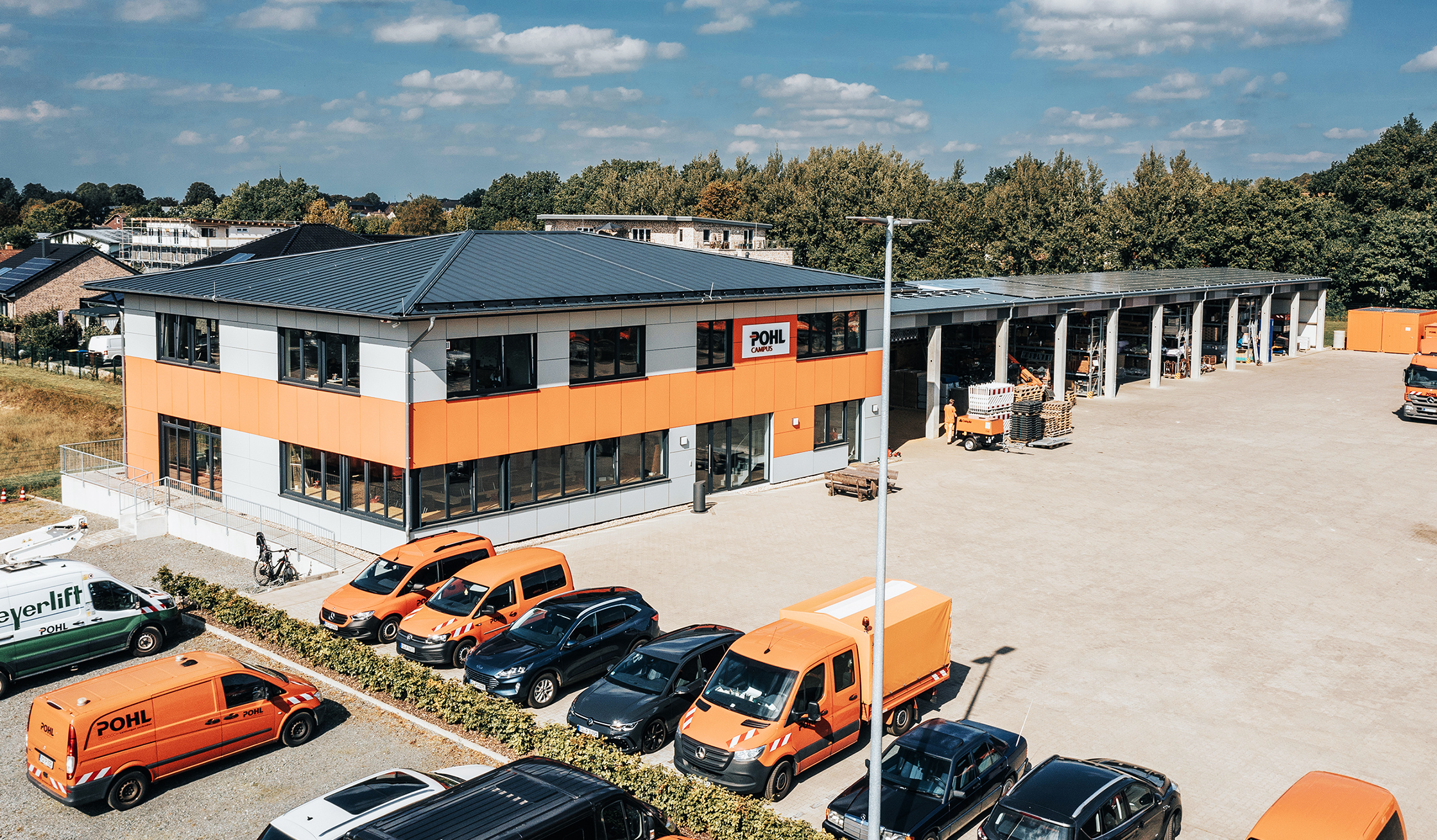 In der Mitte steht ein zweigeschossiges Gebäude mit grauen Fassaden und einem orangefarbenen Band; über dem Eingang hängt das Schild „POHL Campus“. Vorne im Bild parken mehrere orange POHL-Transporter zwischen dunklen Pkw; rechts schließt sich eine lange, offene Lagerhalle mit Regalen und Material an. Im Hintergrund verläuft eine Baumreihe vor blauem Himmel; links sind Nachbargebäude zu sehen.