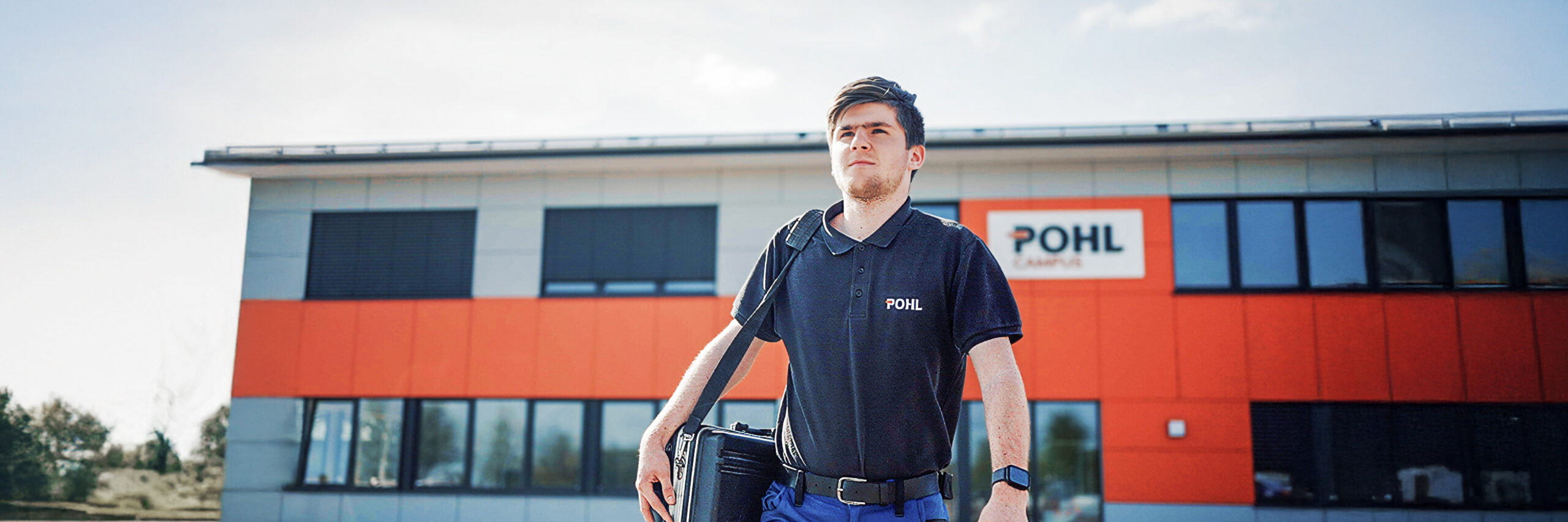 In der Mitte geht eine Person im dunklen POHL-Poloshirt mit einer schwarzen Tasche über den Hof; im Hintergrund steht ein Gebäude mit großem POHL-Schild und orangefarbener Fassade.