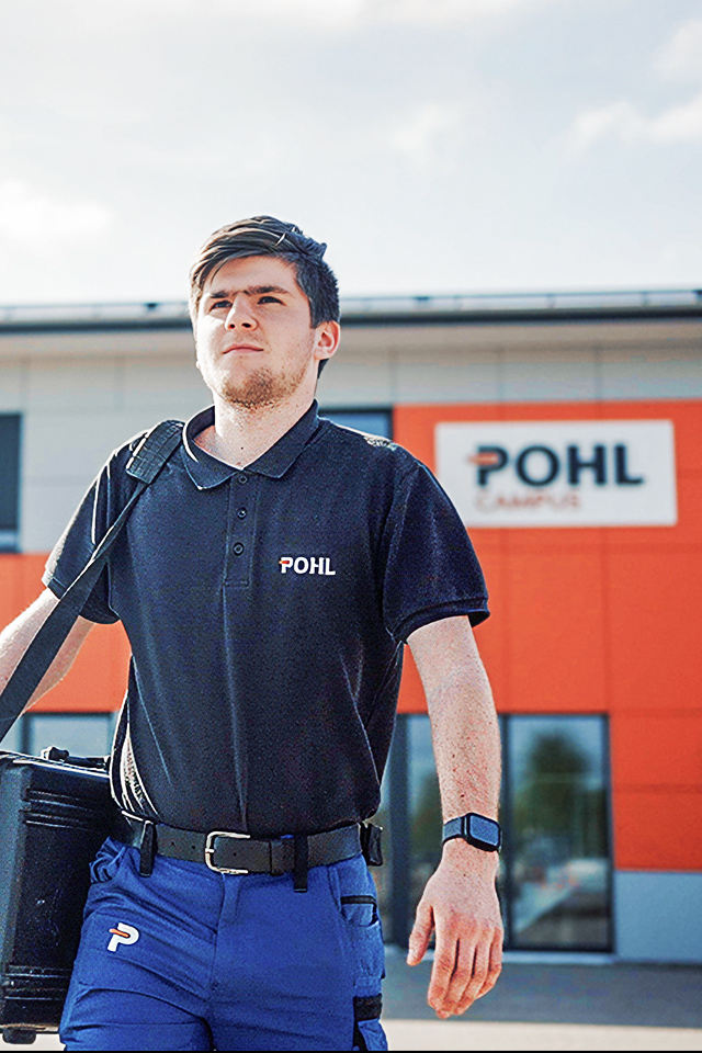 In der Mitte geht eine Person im dunklen POHL-Poloshirt mit einer schwarzen Tasche über den Hof; im Hintergrund steht ein Gebäude mit großem POHL-Schild und orangefarbener Fassade.