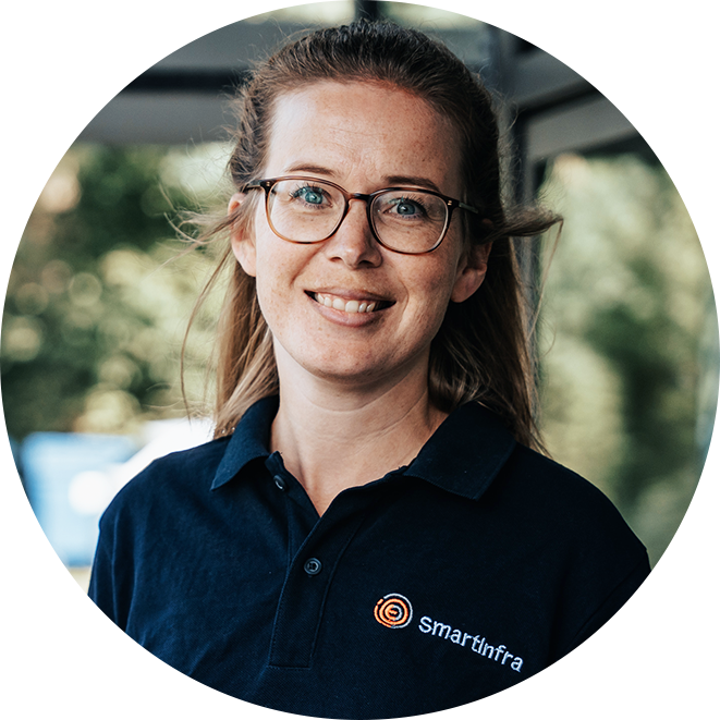 In der Mitte steht eine weibliche Person mit hellbraunen, mittellangen, nach hinten gebundenen Haaren und Brille; sie trägt ein dunkelblaues Poloshirt mit Smartinfra-Logo auf der Brust. Im Hintergrund sind Bäume unscharf erkennbar.