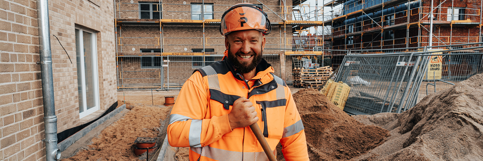 In der Mitte steht ein POHL-Mitarbeiter in orangefarbener Arbeitskleidung und Helm mit einem Stielwerkzeug in der Hand. Links im Bild befindet sich die Fassade eines Neubaus mit Fenstern. Rechts im Bild liegen hohe Sandhaufen und Bauzäune. Im Hintergrund stehen Gerüste an weiteren Gebäuden. Vorne im Bild verläuft ein schmaler, sandiger Arbeitsbereich.