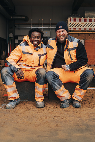 In der Mitte sitzen zwei POHL-Mitarbeiter in orangefarbener Arbeitskleidung nebeneinander auf einem niedrigen Block. Im Hintergrund hängen orangefarbene Trennwände mit sichtbarem POHL-Schriftzug sowie weiße Absperrelemente. Links im Bild stehen Geräte und Rohre auf einer erhöhten Kante. Rechts im Bild stehen große Säcke mit Material. Vorne im Bild liegt ein ebener Sandboden.
