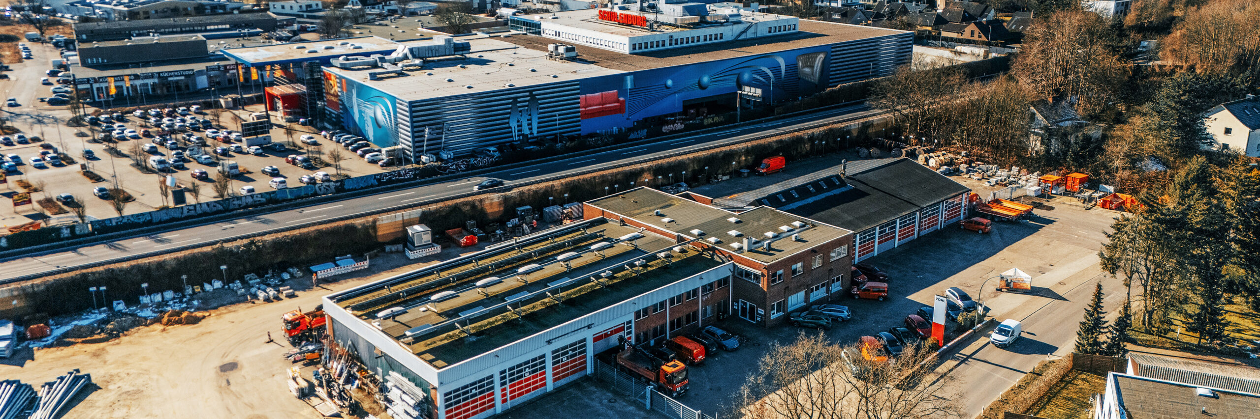Standort Rellingen: Oben links im Bild liegt ein großes Einkaufsgebäude; unten und in der Mitte erstreckt sich ein Betriebshof mit flachen Hallen. Auf dem Hof stehen mehrere orange POHL-Fahrzeuge, rechts hinten sind weitere Abstellflächen mit Containern. Unten rechts verläuft eine Straße; im Hintergrund rechts beginnt ein Wohngebiet mit Bäumen.