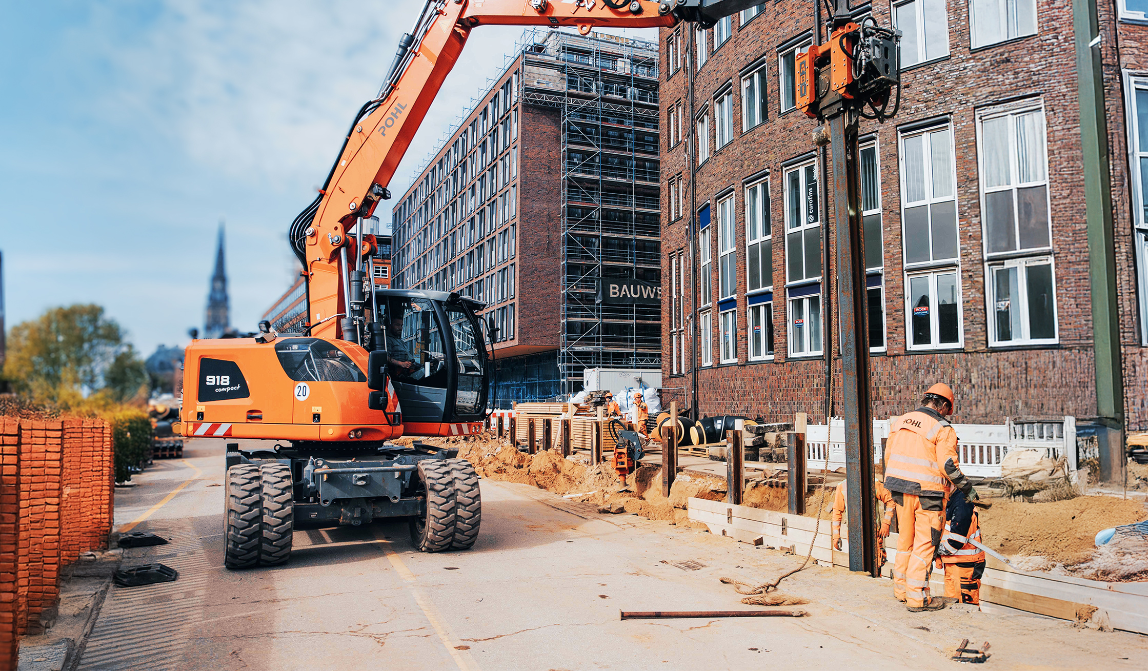 Bagger von POHL auf einer städtischen Baustelle, Mitarbeitende von POHL arbeiten am Gehwegrand.
