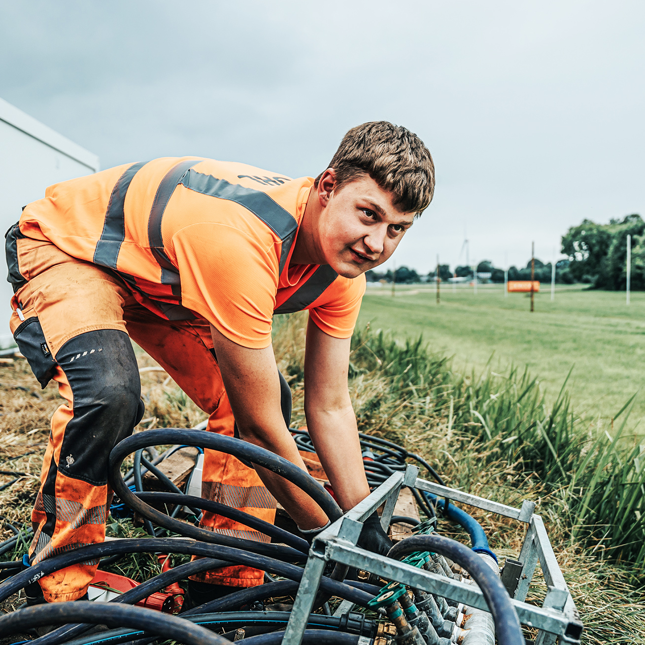 Vorne im Bild kniet ein POHL-Mitarbeiter im Gras und führt mehrere dicke Schläuche in einen Metallverteilerrahmen. Links stehen weiße Container; nach rechts öffnet sich eine lange Wiese mit einzelnen Masten unter bedecktem Himmel.