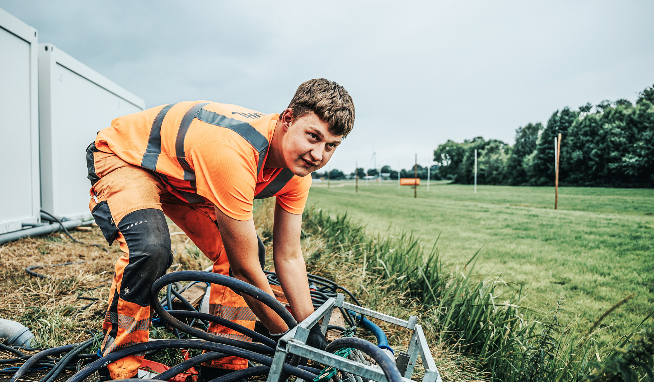 Vorne im Bild kniet ein POHL-Mitarbeiter im Gras und führt mehrere dicke Schläuche in einen Metallverteilerrahmen. Links stehen weiße Container; nach rechts öffnet sich eine lange Wiese mit einzelnen Masten unter bedecktem Himmel.