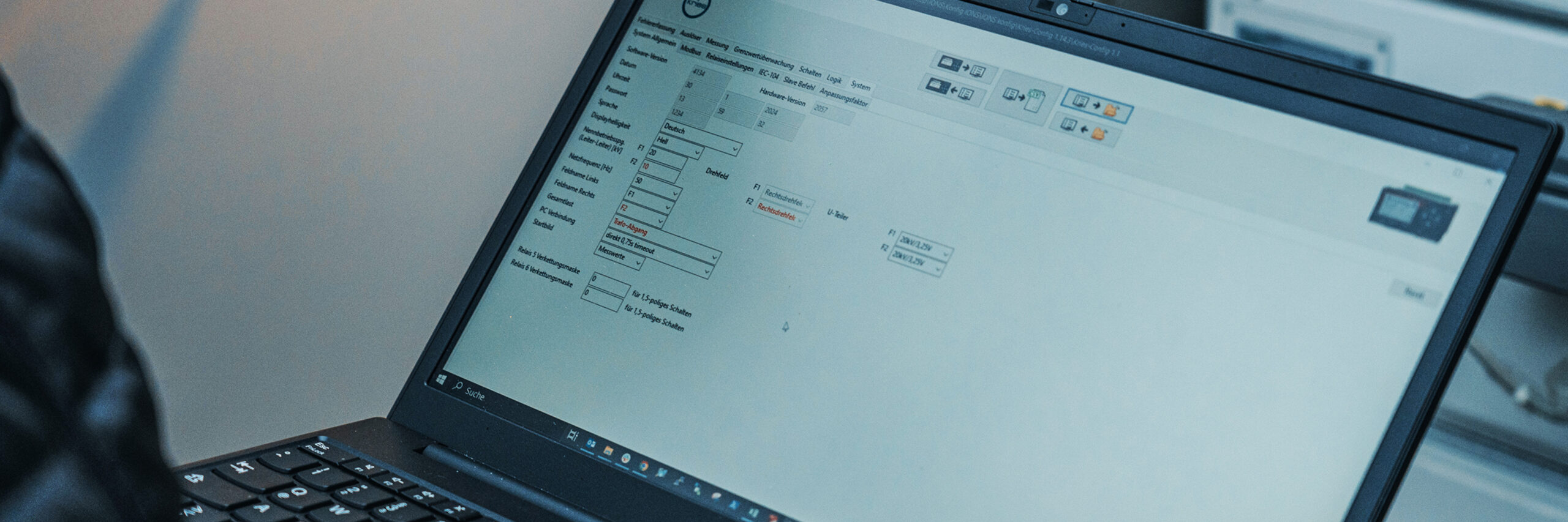Vorne im Bild steht ein aufgeklappter Laptop; auf dem Bildschirm ist eine helle Konfigurationsoberfläche mit Eingabefeldern und Symbolen zu sehen. Rechts im Hintergrund stehen geöffnete Schaltschränke.
