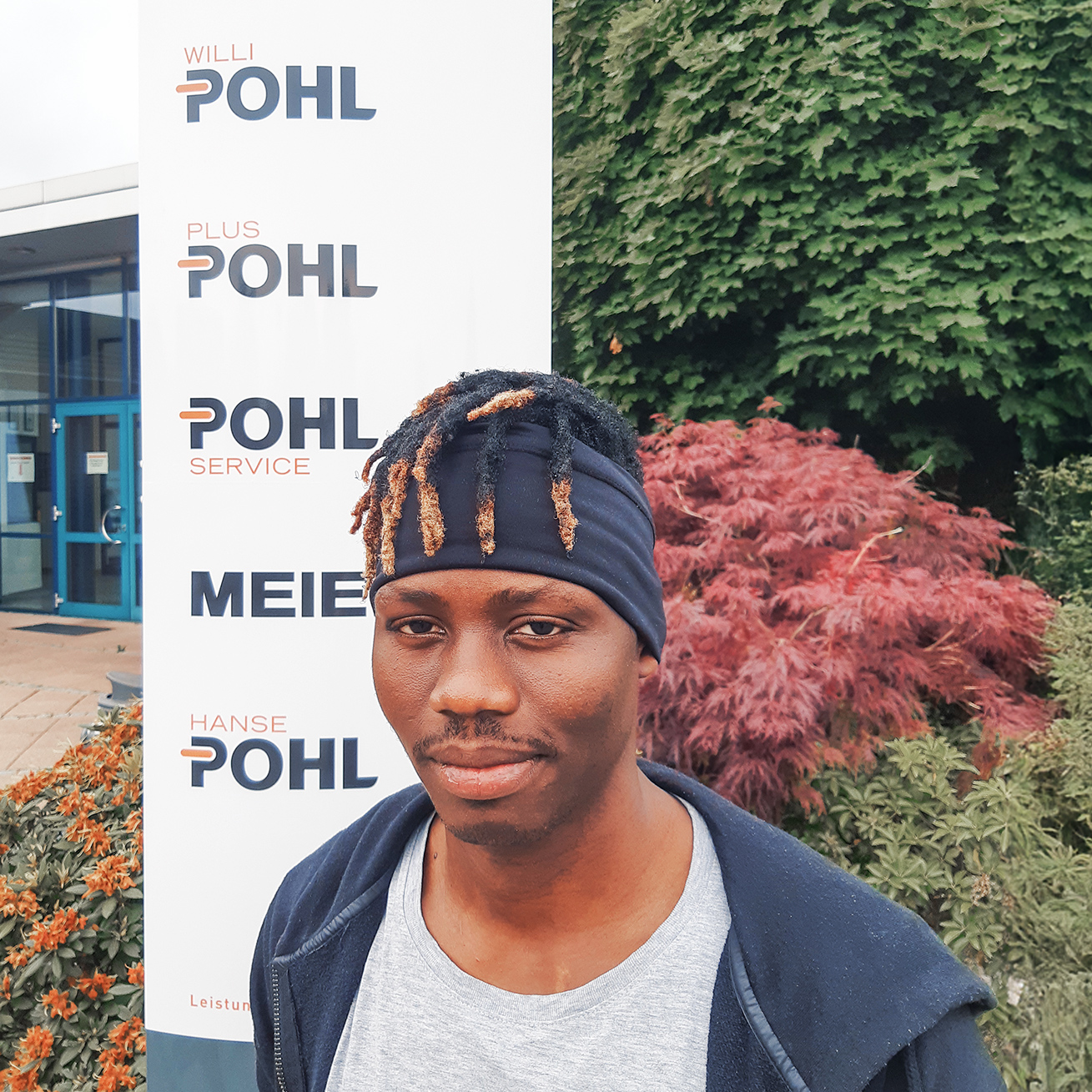 Brustbild, Person mittig-vorn. Dunkles Stirnband mit kurzen, helleren Dreadlocks; dunkle Jacke über grauem T-Shirt. Links im Hintergrund Eingangsbereich mit blauer Glastür, daneben ein weißer Aufsteller mit Firmenlogos. Rechts Sträucher und roter Zierahorn. Bewölktes Tageslicht.