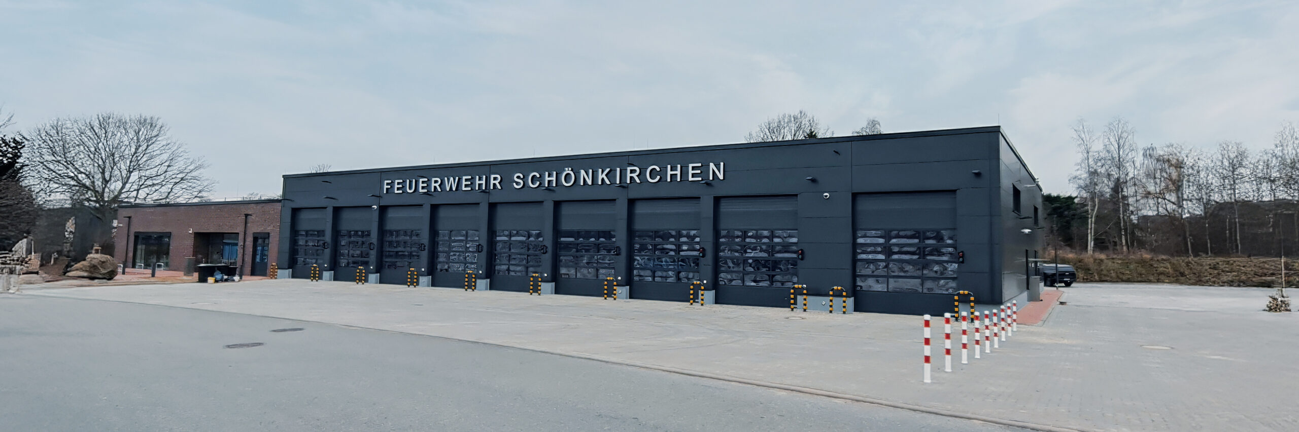 Referenz Neubau Feuerwehr Schönkirchen: Neubau der Feuerwehr Schönkirchen mit dunkler Fassade und mehreren großen Toren für Einsatzfahrzeuge, davor ein gepflasterter Vorplatz.