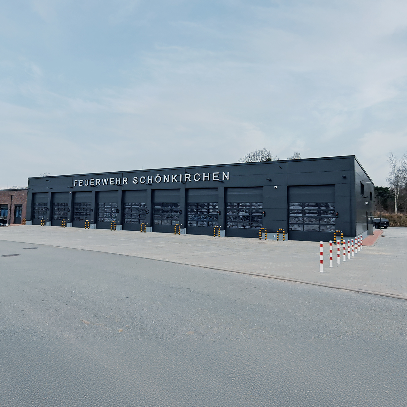 Referenz Neubau Feuerwehr Schönkirchen: Neubau der Feuerwehr Schönkirchen mit dunkler Fassade und mehreren großen Toren für Einsatzfahrzeuge, davor ein gepflasterter Vorplatz.