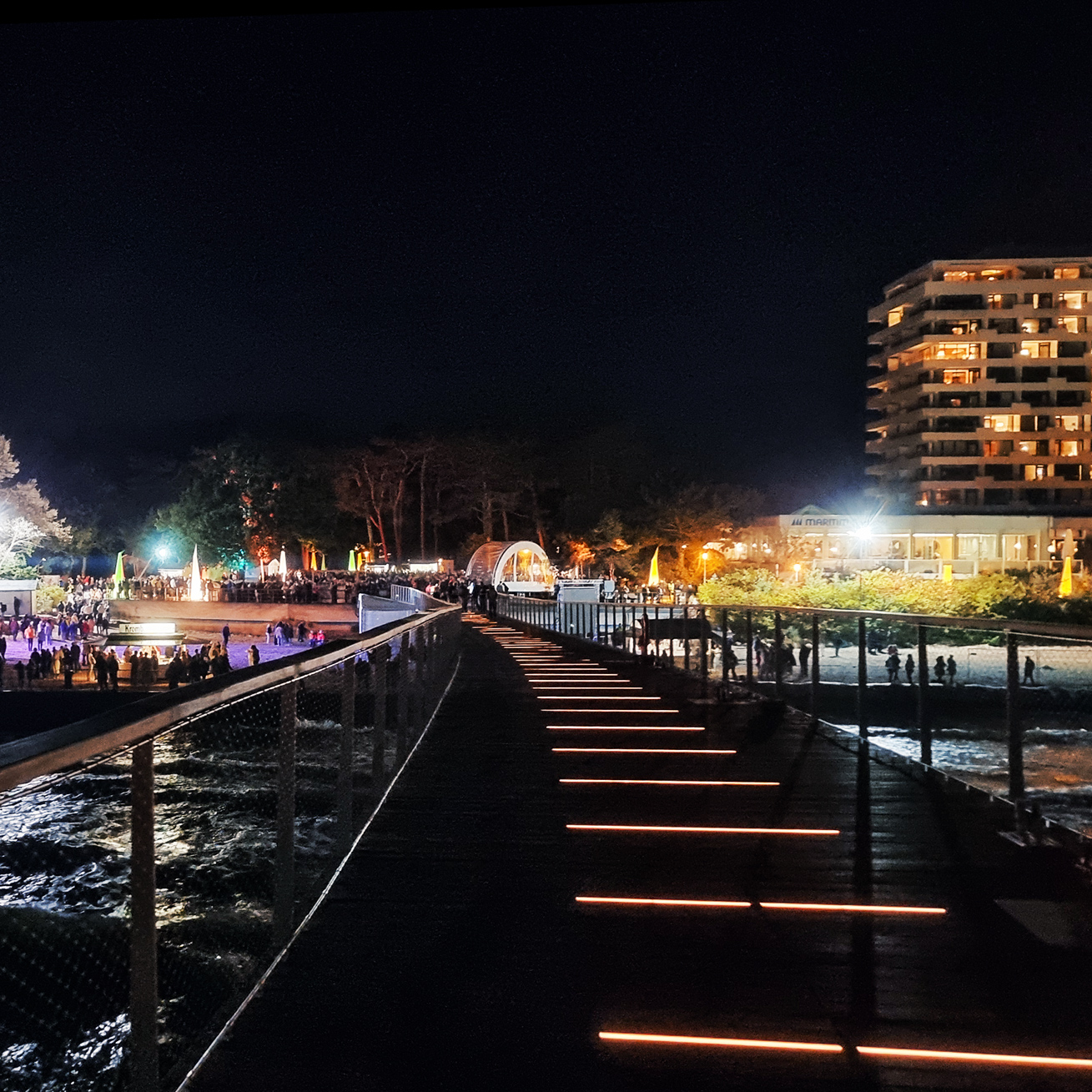 Referenz Maritim Seebrücke Timmendorfer Strand: Von rechts unten nach links oben verläuft ein Steg mit schmalen Leuchtstreifen. Links am Strand drängen sich Menschen vor Bühnen und Ständen; rechts im Hintergrund steht ein hohes Hotelgebäude.