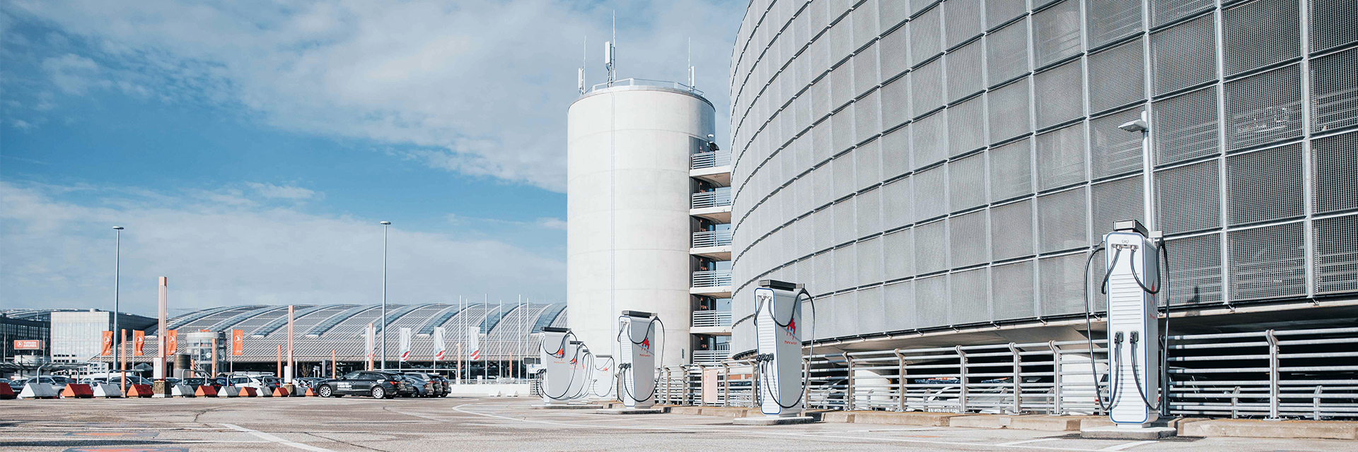 In der Mitte stehen mehrere hohe Ladesäulen auf der Parkfläche eines großen Gebäudekomplexes. Rechts im Bild verläuft eine runde Fassade mit engmaschiger Metallstruktur. Links im Bild sind Parkplätze und weitere Gebäude. Im Hintergrund ragt ein zylindrischer Treppenturm auf. Oben im Bild ist ein teils bewölkter Himmel.