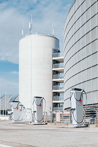 In der Mitte stehen mehrere hohe Ladesäulen auf der Parkfläche eines großen Gebäudekomplexes. Rechts im Bild verläuft eine runde Fassade mit engmaschiger Metallstruktur. Links im Bild sind Parkplätze und weitere Gebäude. Im Hintergrund ragt ein zylindrischer Treppenturm auf. Oben im Bild ist ein teils bewölkter Himmel.