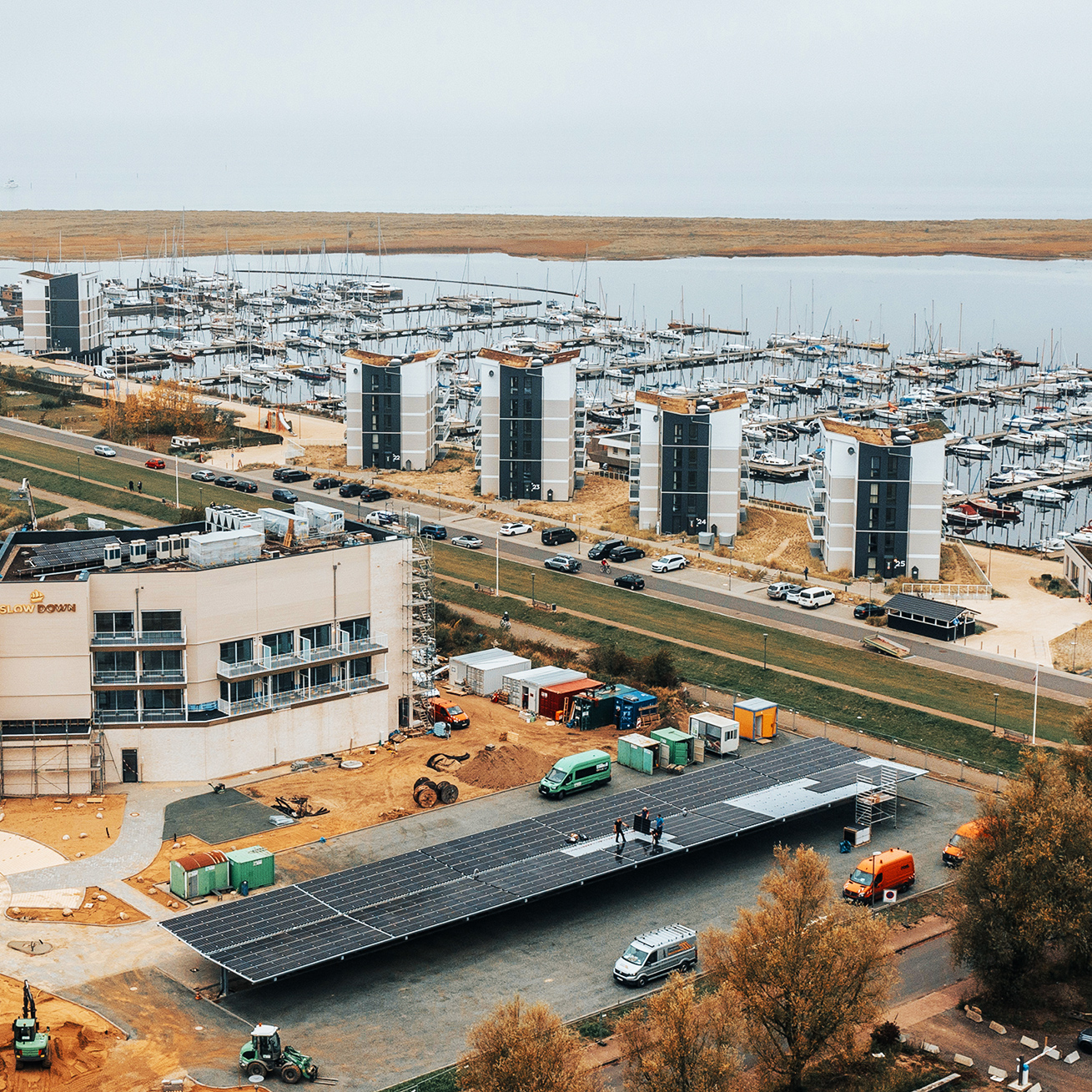 Referenz SlowDown Ostsee-Lagune: In der Mitte stehen mehrere moderne Wohnblöcke an einem Yachthafen mit dicht gereihten Booten. Unten im Bild wird an einem Gebäudekomplex gearbeitet; links liegt ein Flachdach mit Solarmodulen, rechts parken Fahrzeuge, darunter zwei orange POHL-Fahrzeuge.