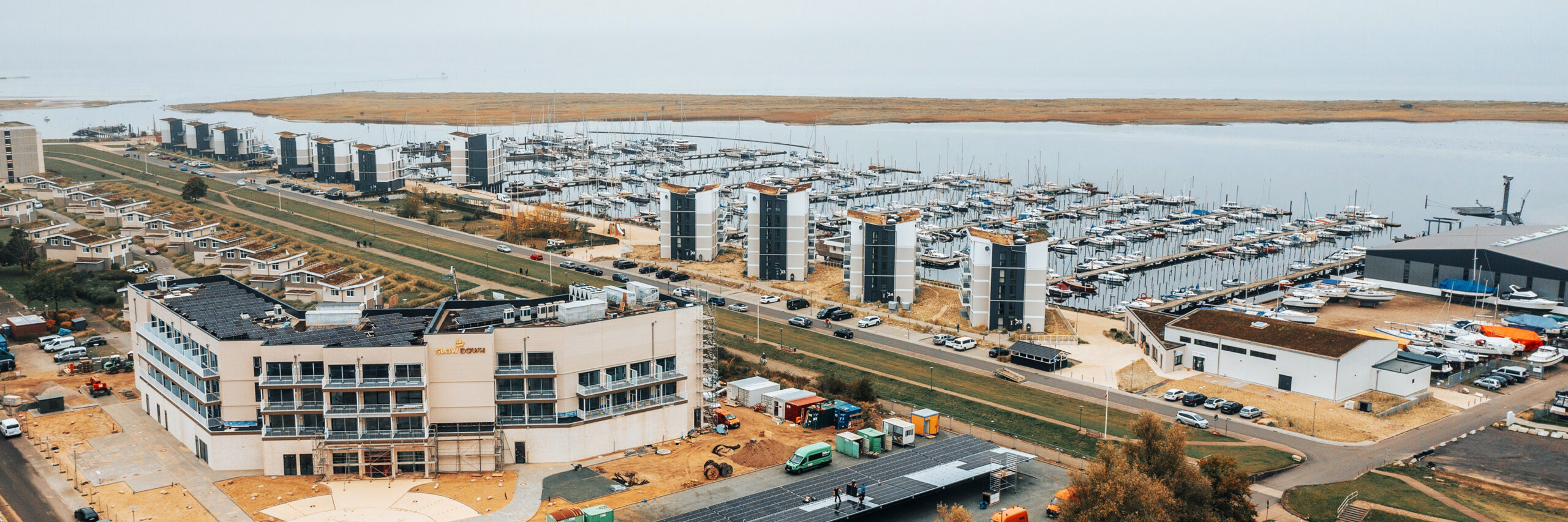 Referenz SlowDown Ostsee-Lagune: In der Mitte stehen mehrere moderne Wohnblöcke an einem Yachthafen mit dicht gereihten Booten. Unten im Bild wird an einem Gebäudekomplex gearbeitet; links liegt ein Flachdach mit Solarmodulen, rechts parken Fahrzeuge, darunter zwei orange POHL-Fahrzeuge.