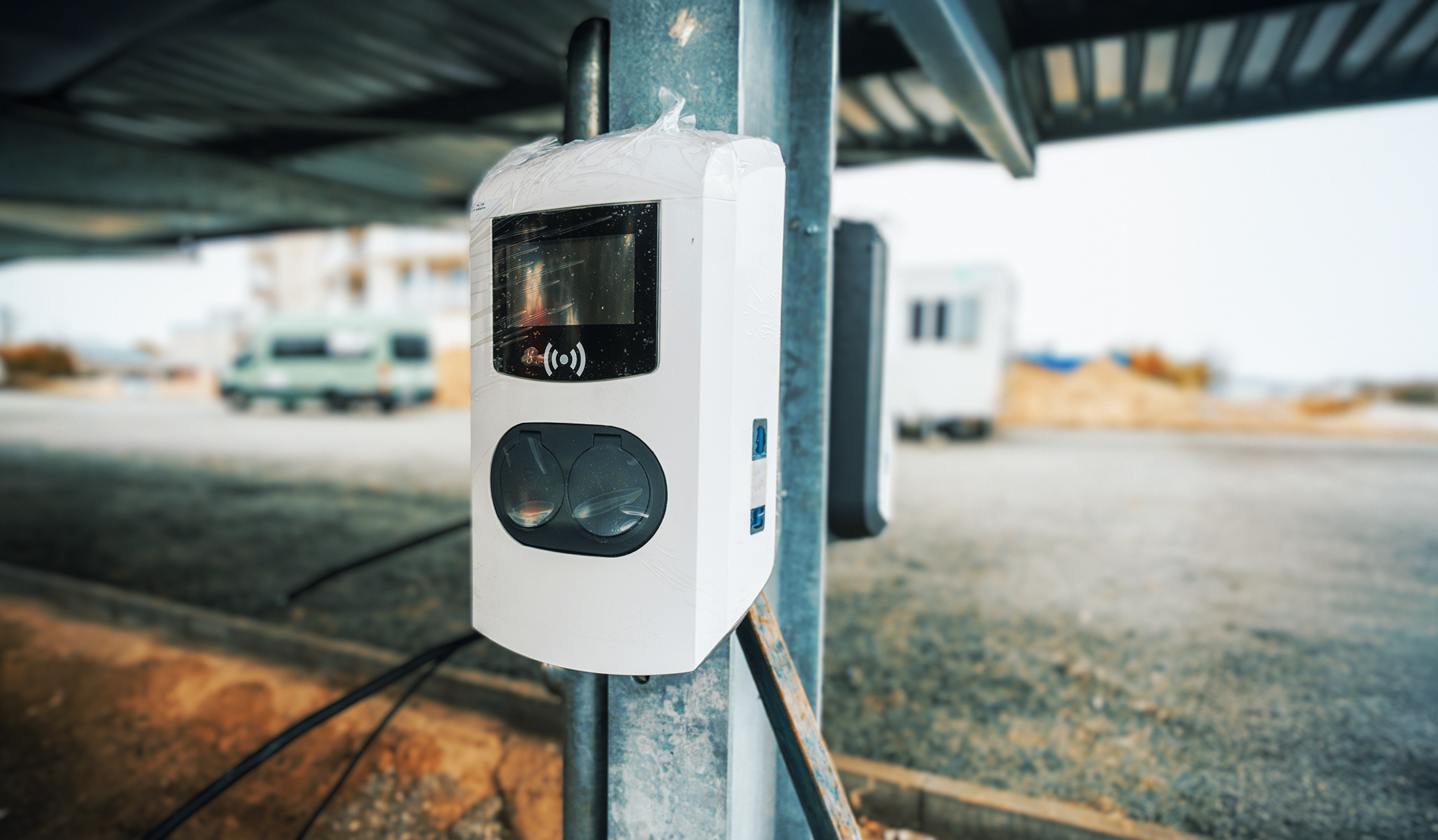 Referenz SlowDown Ostsee-Lagune: Rechts im Bild ist an einem Stahlpfosten ein weißer Kasten mit Display und zwei runden Abdeckungen befestigt. Im Hintergrund unter dem Dach eines Carports stehen Fahrzeuge und Container unscharf; unten liegen lose Kabel.