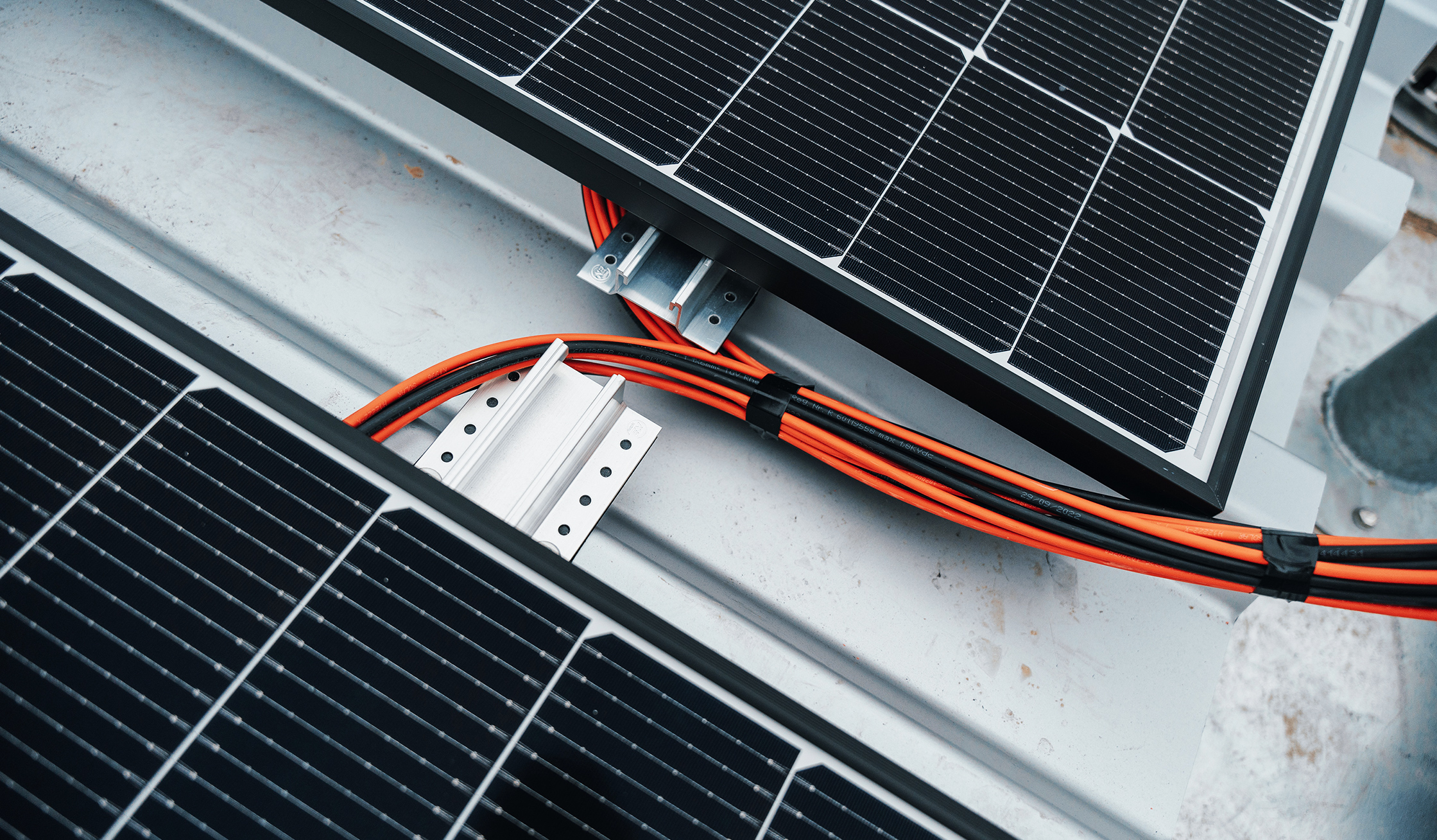 Referenz SlowDown Ostsee-Lagune: Vorne im Bild verlaufen zwei orange Kabelbündel zwischen zwei schwarzen Solarmodulen. In der Mitte halten silberne Schienen die Kabel auf einem grauen Blechdach.
