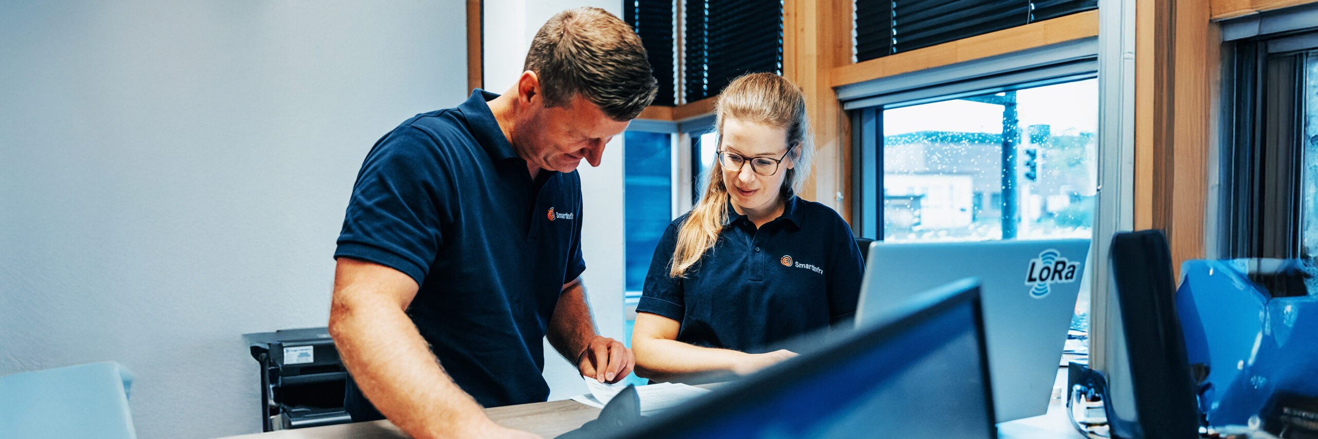 Zwei Mitarbeitende in blauen Poloshirts mit Smartinfra-Logo stehen im Büro. Sie besprechen Dokumente an einem Tisch und wirken konzentriert.