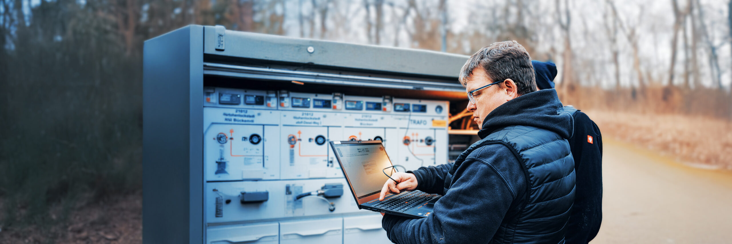 Ein Mitarbeiter mit Laptop steht vor einem großen Schaltkasten im Freien. Er prüft die Technik, während im Hintergrund Kabelsysteme sichtbar sind.