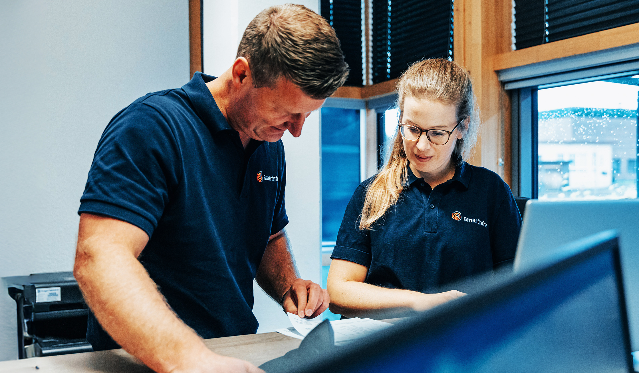 Zwei Mitarbeitende in blauen Poloshirts mit Smartinfra-Logo stehen im Büro. Sie besprechen Dokumente an einem Tisch und wirken konzentriert.