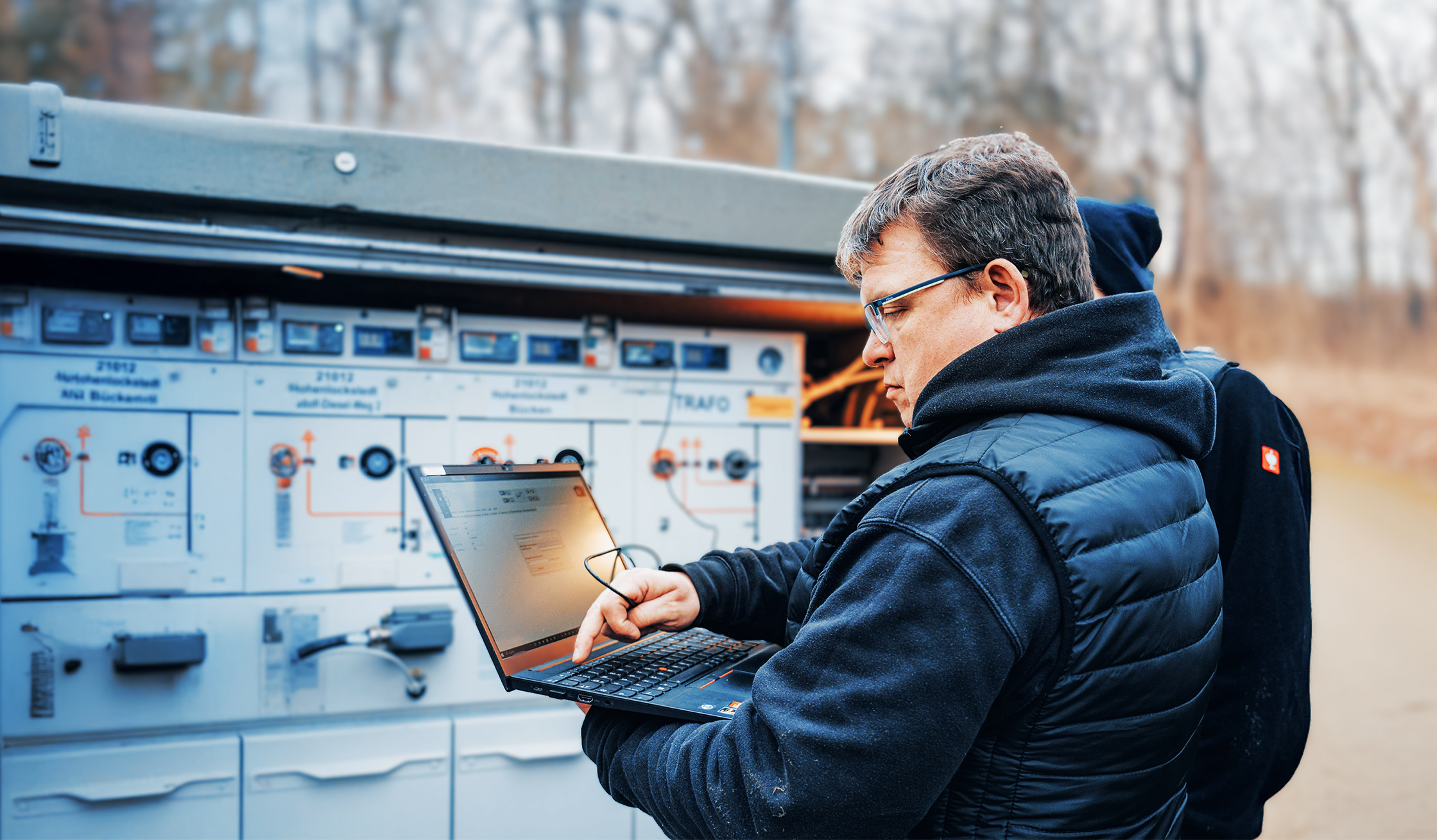 Ein Mitarbeiter mit Laptop steht vor einem großen Schaltkasten im Freien. Er prüft die Technik, während im Hintergrund Kabelsysteme sichtbar sind.