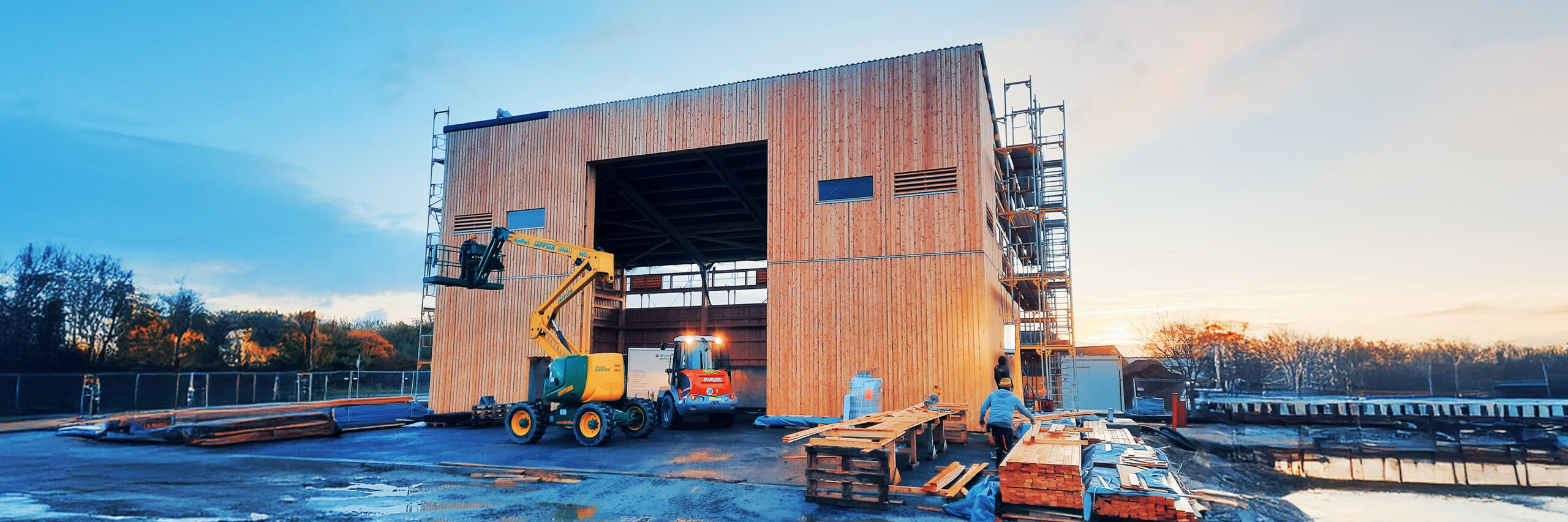 Großes Holzgebäude im Bau mit Baugerüst und Arbeitsfahrzeugen auf der Baustelle.