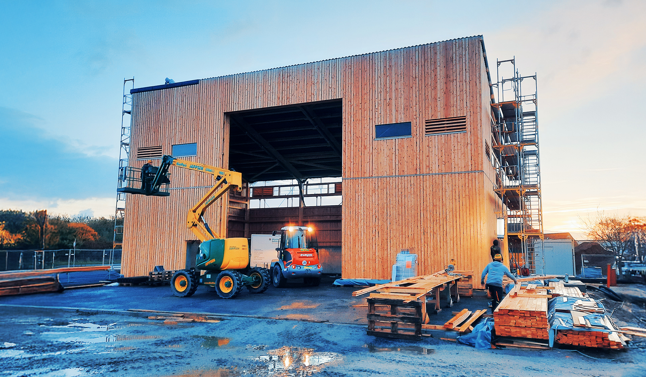 Großes Holzgebäude im Bau mit Baugerüst und Arbeitsfahrzeugen auf der Baustelle.