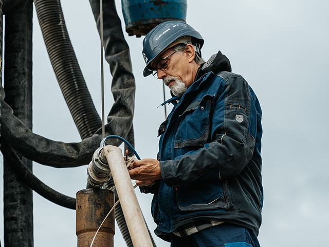 Standort Osterrönfeld: Vorne im Bild arbeitet eine Person mit blauem Helm und dunkler Jacke an dicken Schläuchen. In der Mitte hält die Person ein Kabel an ein Rohrstück. Links und rechts hängen schwarze Schläuche nach unten. Im Hintergrund ist grauer Himmel.