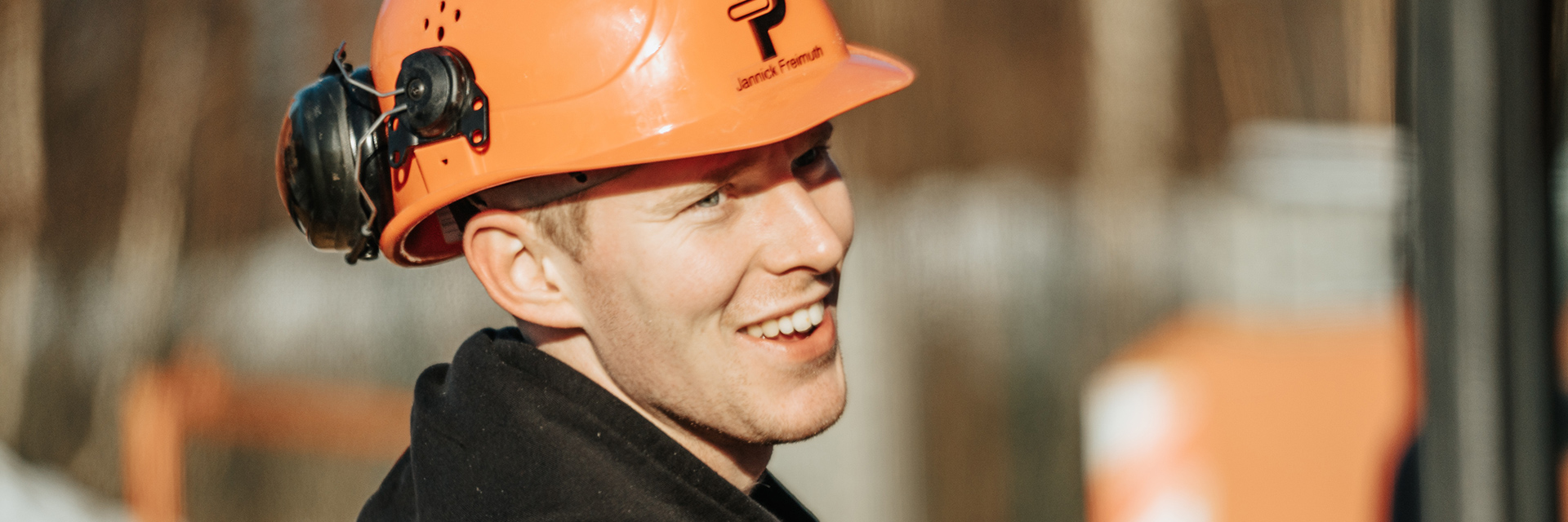 In der Mitte ist der Kopf eines POHL-Mitarbeiters mit orangefarbenem Helm im Profil zu sehen; links am Helm ist Gehörschutz befestigt, im Hintergrund ist alles unscharf.