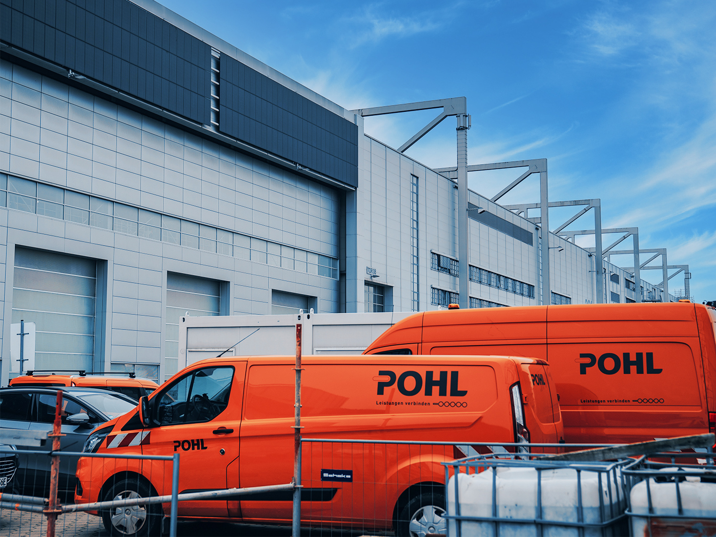Standort Hamburg Finkenwerder: Vorne im Bild stehen mehrere POHL-Fahrzeuge in Orange nebeneinander auf einem Parkplatz. In der Mitte ragt die Seitenwand eines großen Gebäudes mit grauen Fassadenplatten nach oben. Rechts am Rand verläuft ein Zaun. Oben im Bild ist blauer Himmel.
