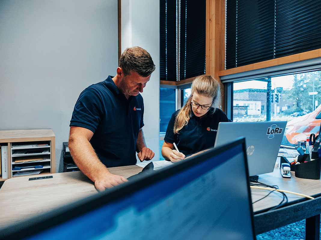 Standort Neumünster: Vorne im Bild liegen Unterlagen auf einem Schreibtisch mit Monitoren. In der Mitte beugen sich zwei Personen über ein Blatt und schreiben. Rechts am Rand steht ein Laptop; draußen vor dem Fenster steht ein POHL-Fahrzeug in Orange. Im Hintergrund sind Regentropfen auf der Scheibe und Bäume.