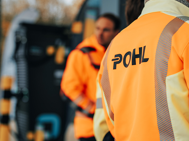 Standort Bützow: Vorne rechts im Bild ist die Rückenpartie eines POHL-Mitarbeiters in orangefarbener Jacke zu sehen; auf der Jacke steht groß „POHL“. Links im Hintergrund stehen weitere POHL-Mitarbeiter unscharf vor Geräten.