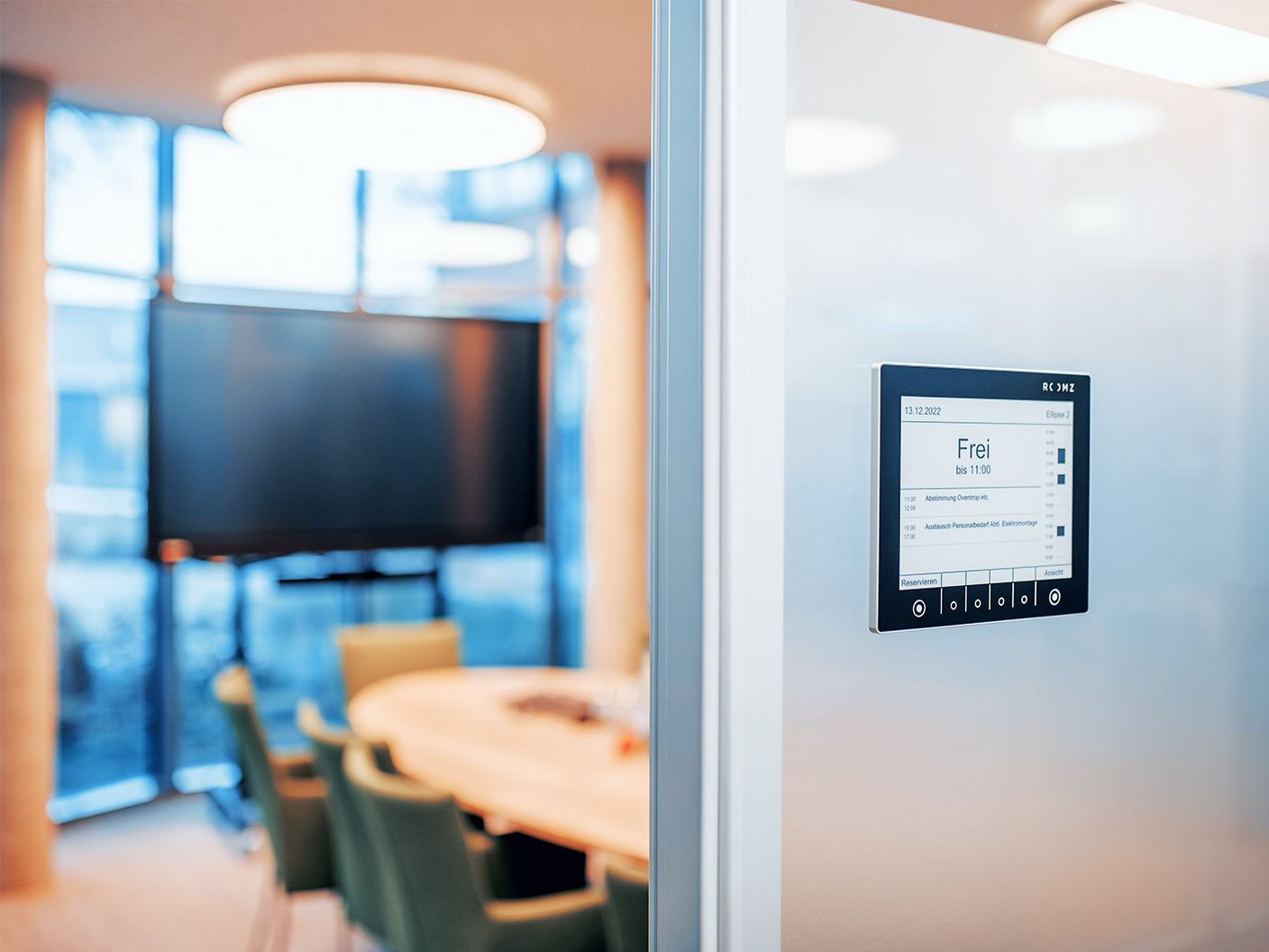 Standort Rendsburg: Rechts im Bild hängt an einer Glaswand ein kleines Display mit Raumbeschriftung; auf dem Display steht „Frei bis 11:00“. Links im Hintergrund steht ein runder Tisch mit grünen Stühlen und ein großer Bildschirm. Oben im Bild leuchten runde Deckenlampen.