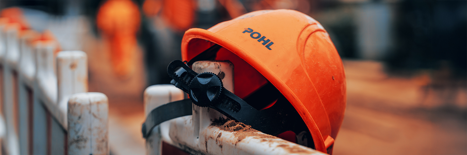 Standort Rendsburg: Vorne im Bild liegt ein orangefarbener Helm mit POHL-Schriftzug auf einem weißen Absperrelement. In der Mitte ist das schwarze Verstellrad des Helms zu sehen. Im Hintergrund verläuft unscharf eine Baustellensituation.