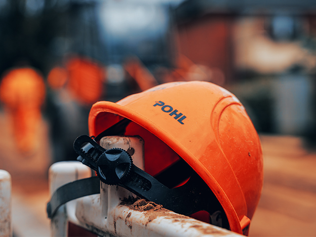 Standort Rendsburg: Vorne im Bild liegt ein orangefarbener Helm mit POHL-Schriftzug auf einem weißen Absperrelement. In der Mitte ist das schwarze Verstellrad des Helms zu sehen. Im Hintergrund verläuft unscharf eine Baustellensituation.