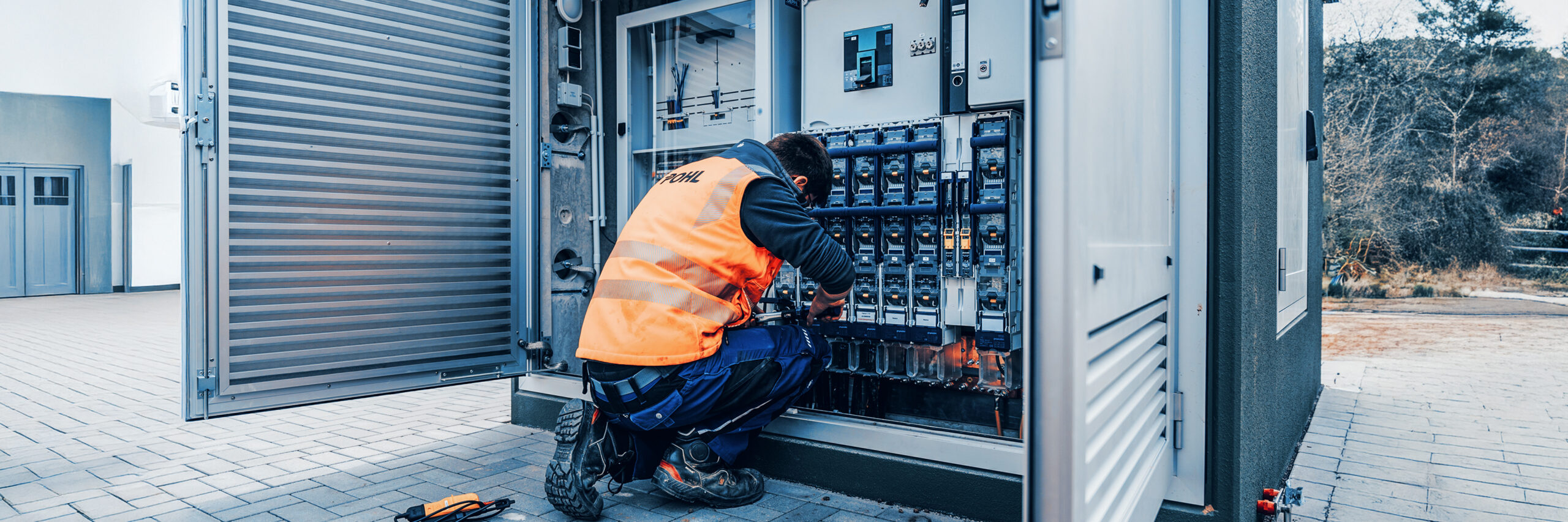 Ein Techniker mit orangener POHL-Warnweste arbeitet im Freien an einem geöffneten Schaltschrank mit vielen Sicherungen und Kabeln.
