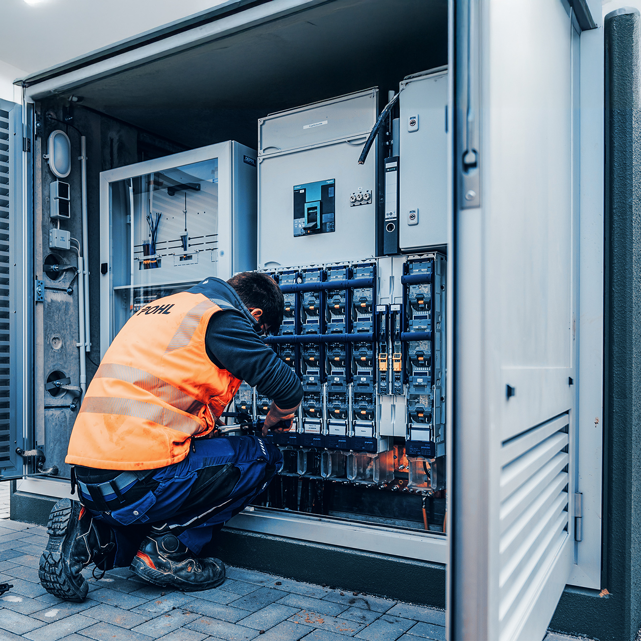 Ein Techniker mit orangener POHL-Warnweste arbeitet im Freien an einem geöffneten Schaltschrank mit vielen Sicherungen und Kabeln.