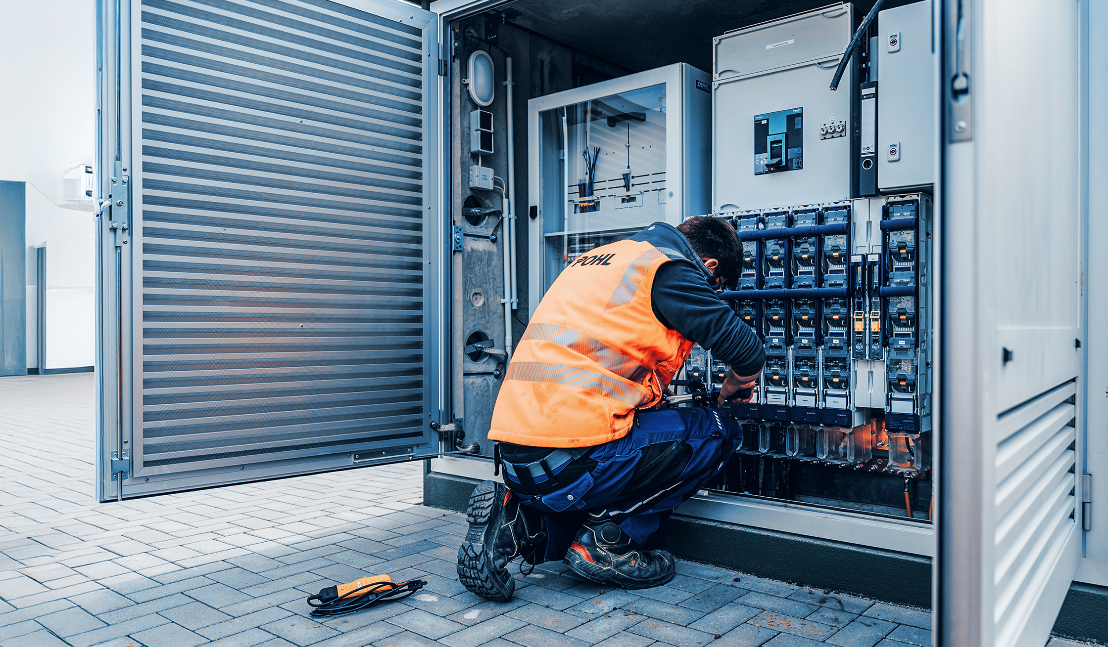 Ein Techniker mit orangener POHL-Warnweste arbeitet im Freien an einem geöffneten Schaltschrank mit vielen Sicherungen und Kabeln.