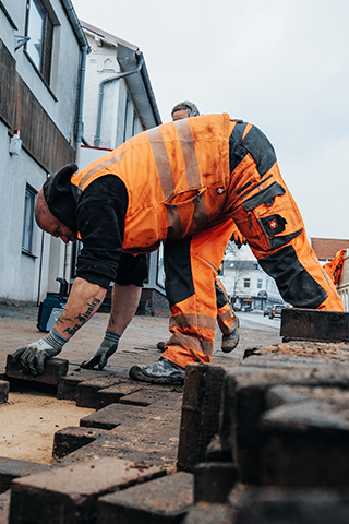 In der Mitte kniet ein POHL-Mitarbeiter in orangefarbener Arbeitskleidung und setzt dunkle Pflastersteine auf einen hellen Sanduntergrund. Links im Bild steht ein weißes Absperrelement und daneben ein Werkzeugkoffer sowie ein handgeführtes Schneidgerät. Rechts im Bild verläuft eine Straße mit geparkten Fahrzeugen, darunter ein orangefarbenes POHL-Fahrzeug. Im Hintergrund stehen Wohn- und Geschäftshäuser. Vorne im Bild liegen geordnete Reihen von Pflastersteinen.