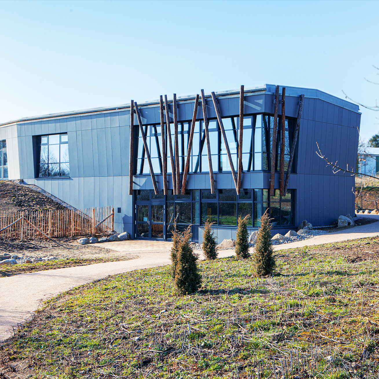 Referenz Steinzeithaus Albersdorf: Modernes Ausstellungsgebäude mit grauer Fassade und großen Glasfronten, davor auffällige Holzstämme als Gestaltungselemente.