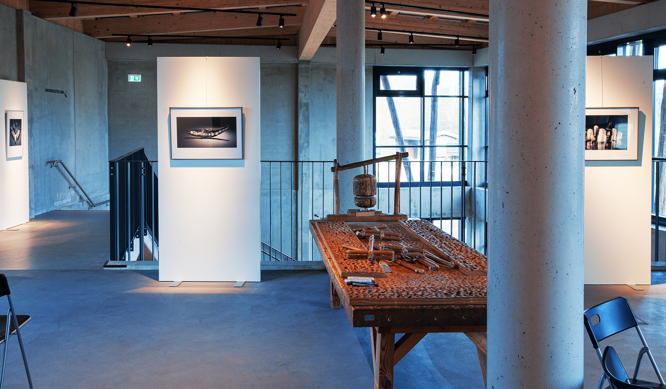 Referenz Steinzeithaus Albersdorf: Innenraum einer Ausstellung mit hellen Wänden und Fotos. In der Mitte steht ein alter Holztisch mit historischen Werkzeugen.